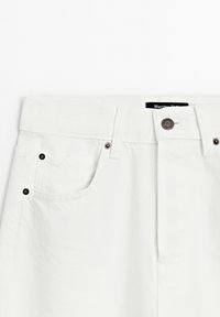 Section de taille d'un jean en denim blanc avec poche avant, passants pour ceinture, bouton en métal et partie de l'étiquette visible à l'intérieur de la ceinture.