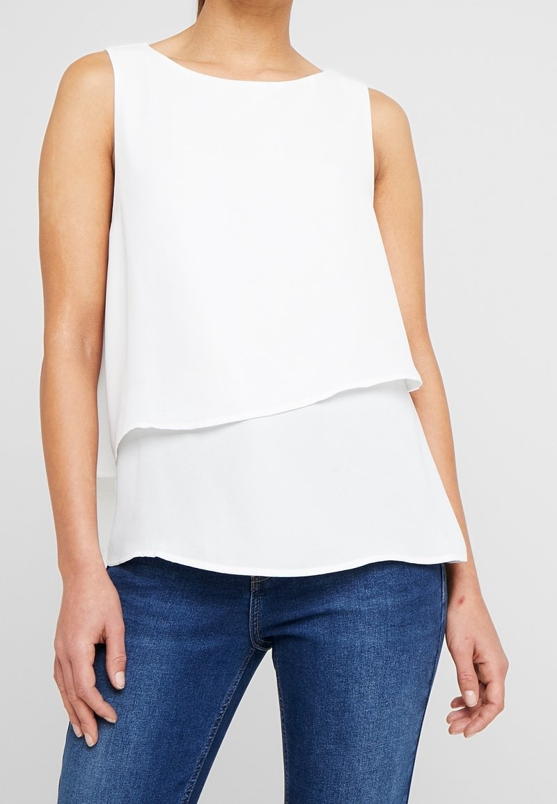 Haut blanc sans manches avec un col rond et un design à couches. Texture lisse, ourlet arrière légèrement plus long, associé à un jean bleu foncé.