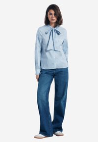 Camicia a righe blu e bianche con dettaglio del papillon, abbinata a pantaloni in denim a gamba larga e scarpe bianche con lacci.