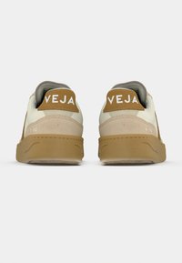Ζευγάρι μπεζ και καφέ αθλητικά παπούτσια Veja V-90, εμφανιζόμενα από πίσω, με λαστιχένιες σόλες και λογότυπο στις πίσω ετικέτες.