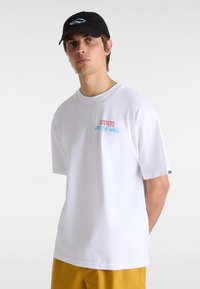 Weißes Baumwoll-T-Shirt mit kurzen Ärmeln, versehen mit dem roten und blauen "VANS OFF THE WALL"-Logo auf der Brust. Abgerundet durch eine schwarze Mütze.