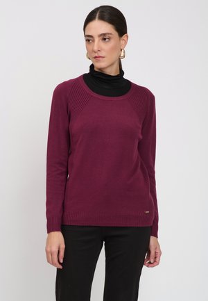 Donna che indossa un maglione viola a maniche lunghe sopra un dolcevita nero, con pantaloni neri, in piedi davanti a uno sfondo chiaro e semplice.