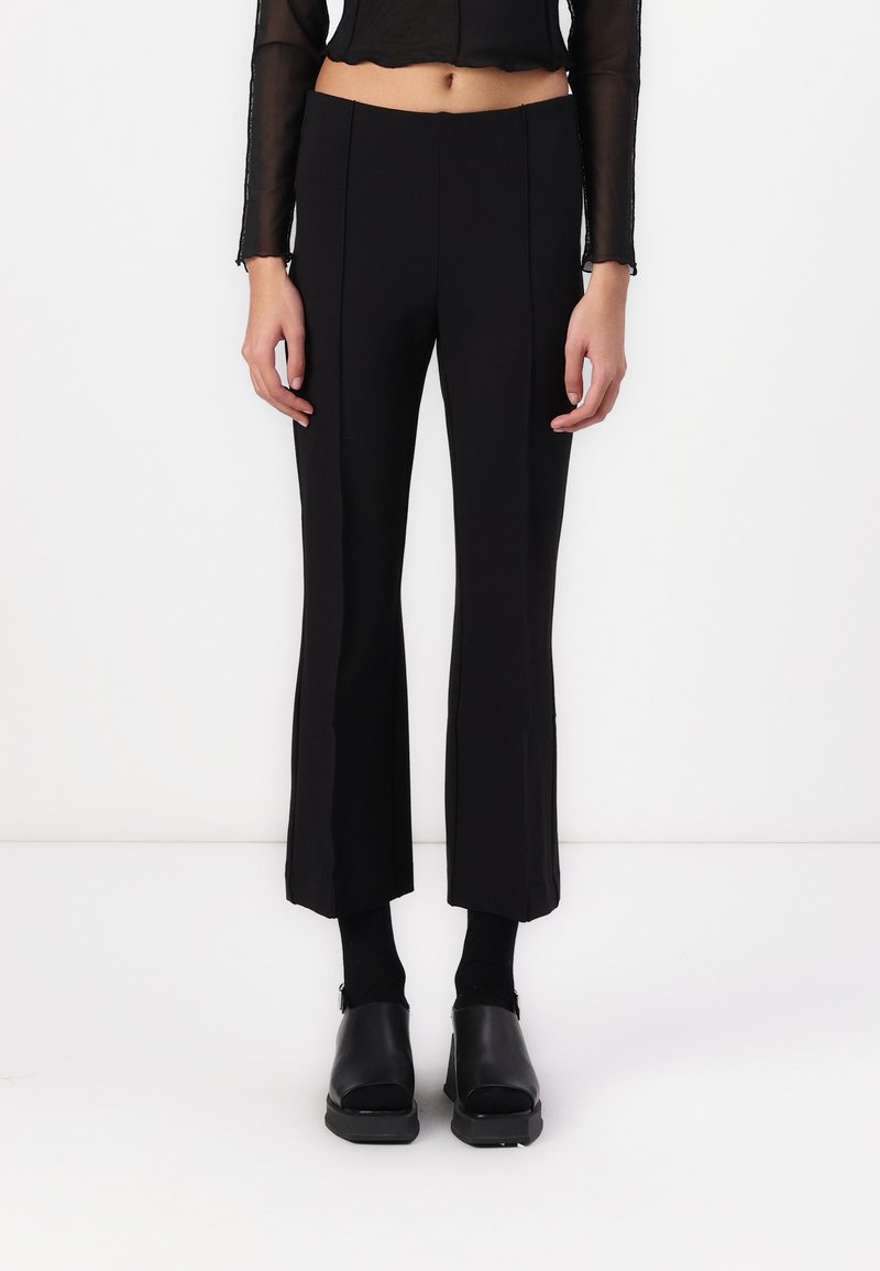 Pantalons évasés noirs et ajustés avec une texture lisse, présentant des coutures verticales sur le devant. Portés avec des chaussettes et des chaussures noires.