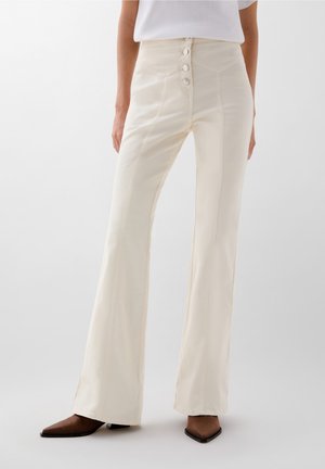 Femme portant un pantalon évasé crème à taille haute avec des boutons à l'avant et des chaussures à bout pointu marron, se tenant contre un fond uni.