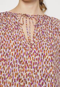 Blouse multicolore avec un col en V et une fermeture à nouer, fabriquée en tissu léger, présentant des motifs abstraits en orange, violet et marron.