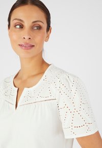 Blusa bianca con scollo rotondo e maniche corte a sbuffo, con motivi ricamati a taglio sulle spalle e sul petto. Tessuto morbido.