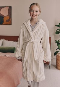 Peignoir blanc côtelé en matière polaire douce avec capuche, fermeture à nouer à la taille et poches latérales, porté par-dessus une chemise légère.