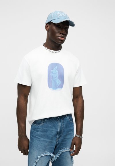 Άνδρας που φοράει ξεβαμμένα ανοιχτόμπλε τζιν, λευκό t-shirt με μπλε γραφικό άγαλμα, μπλε καπέλο μπέιζμπολ, ασημένιο αλυσίδα-κολιέ και βραχιόλι μπροστά σε λευκό φόντο.