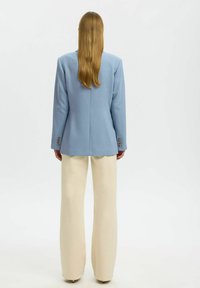 Femme aux longs cheveux blonds portant un blazer bleu clair et un pantalon ample crème, debout de dos sur un fond uni.