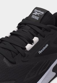 Μαύρο αθλητικό παπούτσι προπόνησης Reebok με μαύρα κορδόνια, λευκές οπές και λευκό λογότυπο στη γλώσσα και στο πλάι, εμφανιζόμενο σε κοντινή γωνία.