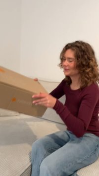 Bruine kartonnen doos met een oranje logo, vastgehouden door een persoon in een kastanjebruine langemouwenshirt, zittend op een lichtgekleurde bank.