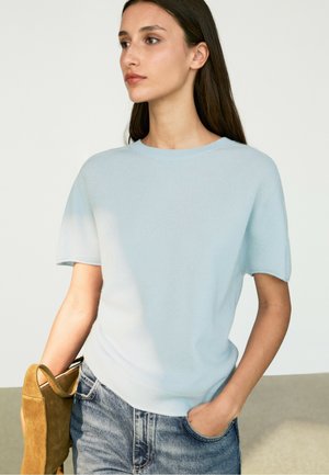 Donna con top azzurro chiaro a maniche corte e jeans blu sbiaditi, che tiene una borsa in camoscio marrone nella mano sinistra, mano destra in tasca, guarda verso sinistra.