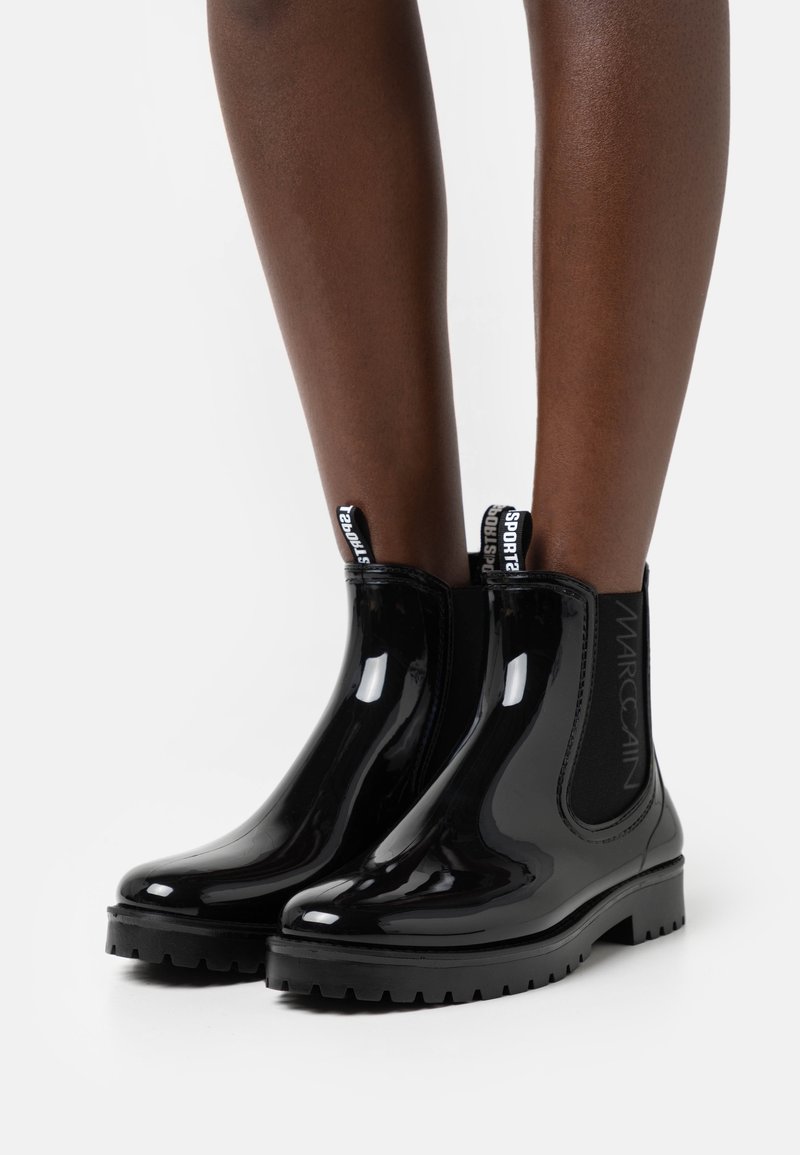 Bottines Chelsea noires et brillantes avec des panneaux élastiques sur les côtés et des languettes. Semelle épaisse avec un relief texturé. Logo de la marque visible sur le côté.