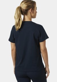 Mujer con cola de caballo rubia vista de espaldas, vestida con una camiseta lisa de manga corta color azul marino oscuro y pantalones a juego, sobre un fondo blanco.