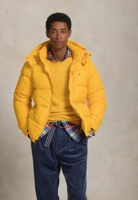 Žuta pufer jakna s kapuljačom, prešivena tekstura, nošena preko žutog duksa, slojevito uz kariranu košulju i tamnoplave hlače.