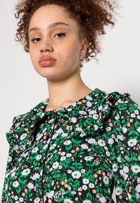 Blusa preta com padrão floral e gola com folhos, apresentando flores verdes, rosas e azuis, e detalhe de botões na gola.