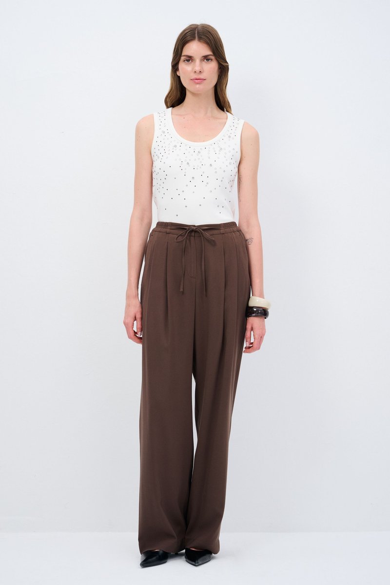 Witte mouwloze top met decoratieve studs bij de halslijn, gecombineerd met losse bruine broek met hoge taille, voorzien van een trekkoord en wijde benen.