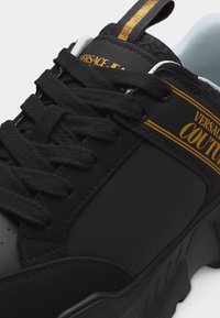 Μαύρο αθλητικό παπούτσι με χοντρές κορδόνια, χρυσή επιγραφή "Versace Couture" στην πλευρά και τη γλώσσα, και ορατό υφή στην άκρη της σόλας.