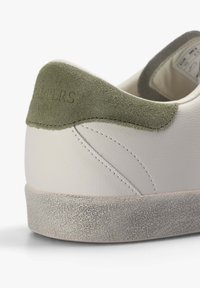 Zapatilla blanca de cuero con una parche de ante verde en el talón y suela gris texturizada. Presenta detalles de costura y un logotipo en relieve en el talón.