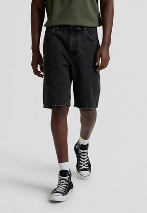 Άνδρας που περπατά φορώντας μαύρα σορτς από τζιν, μαύρα ψηλά αθλητικά παπούτσια με λευκά κορδόνια, λευκές κάλτσες και πουκάμισο χακί.