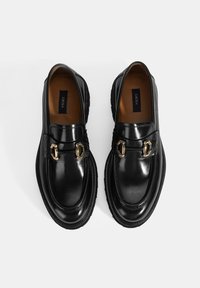 Zwarte leren loafers met een glanzende afwerking, voorzien van gouden hardware-accenten en een dikke rubberen zool. Binnenvoering van lichtbruin materiaal.