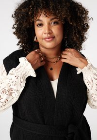 Zwart gevoerde omslag met een strik, gelaagd over een witte top met kant uitsparingen op de mouwen. Gouden kettingen en hangerkettingen worden gedragen.