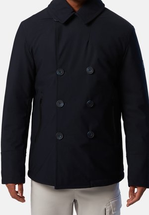 Mann trägt eine schwarze, doppelreihige Jacke mit breitem Kragen und seitlichen Reißverschlusstaschen, kombiniert mit hellfarbenen Cargohosen.