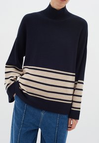 Pulover tricotat de culoare bleumarin cu guler înalt, având dungi crem și închise, croială lejeră și textură cu nervuri, asortat cu blugi blue.