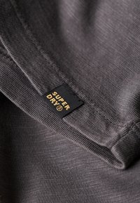 Tejido gris con una superficie texturizada; incluye una etiqueta negra con el nombre de la marca "SUPER DRY" en dorado. Vista de cerca del borde del material.