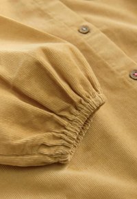 Tissu en velours côtelé jaune moutarde avec un ourlet de manches froncé et des détails de boutons, présentant une texture côtelée subtile et une finition douce.