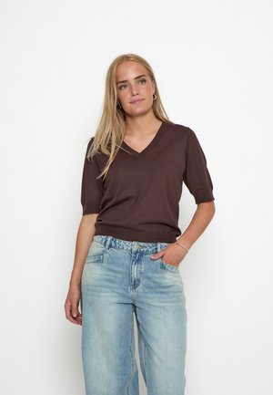 Blond haar vrouw die een bruine korte mouw V-hals trui draagt en lichtblauwe hoog getailleerde spijkerbroek met één hand in de zak tegen een witte achtergrond.