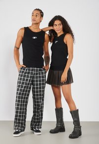 Deux mannequins portent des débardeurs noirs. L'un des mannequins porte un pantalon ample à carreaux, tandis que l'autre porte une jupe noire et des bottes grises hautes avec des accents métalliques.