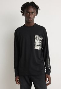 Zwarte longsleeve shirt met een gemêleerd wit grafisch ontwerp op het voorzakgebied en opdruk langs de linkermouw. De stof lijkt glad.