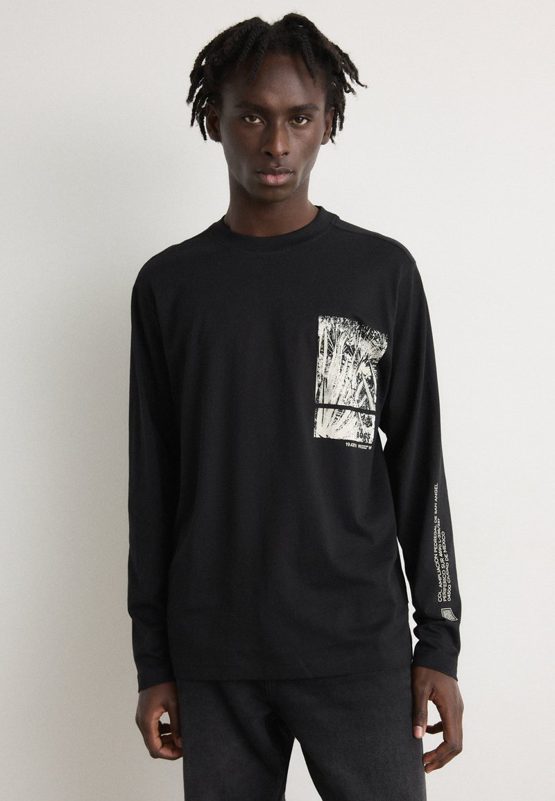 Black long-sleeve shirt with a textured white graphic on the front pocket area and printed text along the left sleeve. Fabric appears smooth.