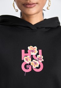 Sudadera negra con un gran logo rosa "HUGO", adornado con gráficos de flores blancas y amarillas. El material parece suave; el diseño es informal y vibrante.