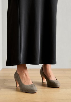 Chaussures à talons hauts en daim gris à bout rond et à talon de hauteur moyenne, accompagnées d'une jupe noire longue sur un sol en bois.