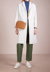 Stribet lysjakke med blå lodrette striber, grønne bukser, hvide sneakers og en tan crossover taske. Glatte teksturer og skræddersyet pasform.