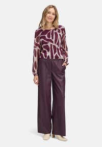 Kobieta ubrana w fioletową wzorzystą, przezroczystą bluzkę z długim rękawem, szerokie ciemnofioletowe spodnie i beżowe botki na obcasie, stojąca na białym tle.
