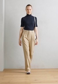 Un col roulé noir à manches courtes, un pantalon taille haute beige et des ballerines grises. Un design simple avec une coupe ajustée, présenté sur un mannequin devant un fond neutre.