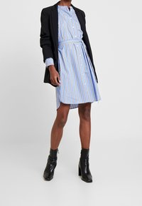 Robe chemise rayée bleue et blanche avec un col, des boutons sur le devant et une taille ceinturée, associée à un blazer noir et des bottines.