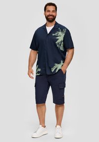Marineblaues Kurzarmhemd mit Knöpfen und grünem Palmenblattmuster auf der linken Seite, kombiniert mit cargo Shorts und weißen Sneakern.
