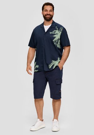 Chemise à manches courtes navy avec imprimé de feuilles de palmier vertes sur le côté gauche, assortie à un short cargo et des baskets blanches.
