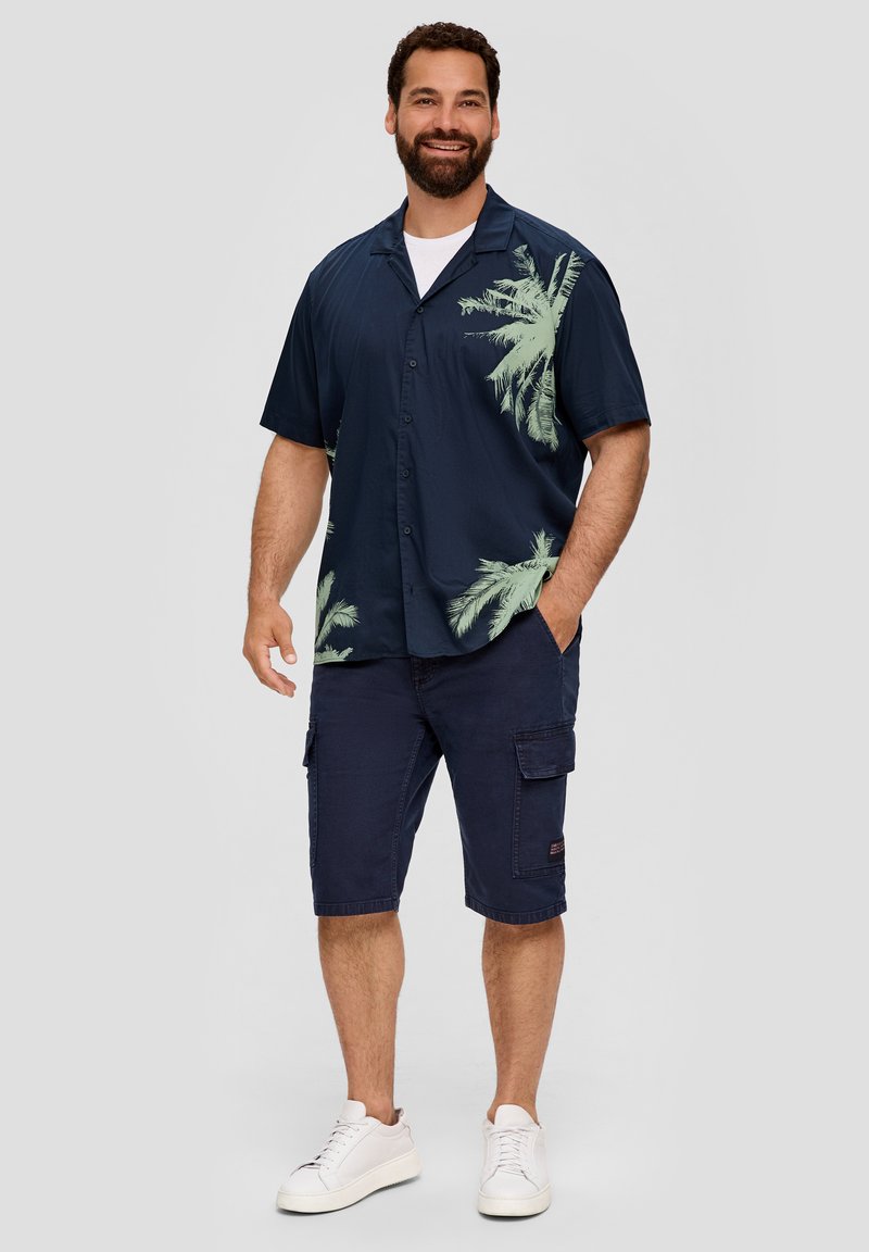 Marineblaues Kurzarmhemd mit Knöpfen und grünem Palmenblattmuster auf der linken Seite, kombiniert mit cargo Shorts und weißen Sneakern.