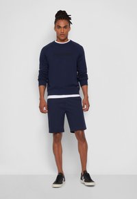 Námořnický crewneck svetr s černým logem "Dockers", doplněný námořnickými šortkami. Model má na sobě černé tenisky. Hladká tkanina, ležérní design.
