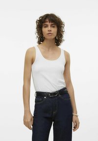 Witte mouwloze tanktop met een ronde halslijn, gecombineerd met een donkere spijkerbroek en een zwarte riem; model heeft krullend haar en draagt een armband.