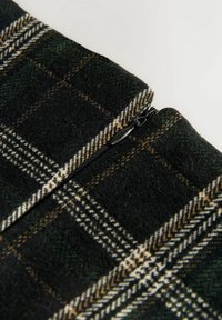 Gros plan sur un tissu à carreaux verts et jaunes avec une fermeture éclair noire partiellement fermée sur un vêtement, posé sur un fond uni clair.