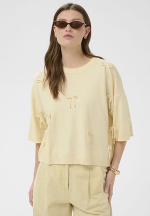 Kobieta w beżowym, oversize'owym swetrze z małymi kokardkami, beżowych spodniach z wysokim stanem, brązowych okularach przeciwsłonecznych w kocim kształcie i złotej biżuterii.