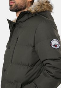 Gevoerde donkergroene winterjas met een met bont afgewerkte capuchon, een ritssluiting op de borstzak en een ronde logopatch op de mouw.