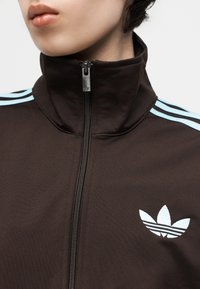 Κοντινό πλάνο ενός ατόμου που φοράει σκούρο καφέ μπουφάν Adidas με ανοιχτό μπλε ρίγες στους ώμους και λευκό λογότυπο Adidas στο στήθος.