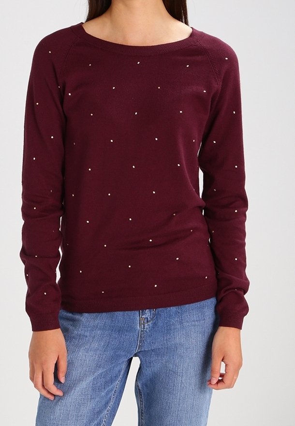Femme portant un pull manches longues bordeaux à petits pois blancs, associée à un jean bleu, debout devant un fond uni.